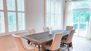 A dining area with a wooden table and six upholstered chairs, large windows with blinds, and a view of greenery outside. - Grande at Canal Pointe home with pools, sports courts, & gym (Rehoboth Beach)