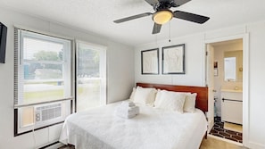 Une chambre à coucher avec un lit, un ventilateur de plafond, des œuvres d’art, et une salle de bain visible à travers une porte ouverte.