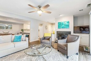 A modern living room with a ceiling fan, a glass coffee table, a sofa, two armchairs, a fireplace, and a TV. - Eagles Landing - updated condo w/two balconies, WiFi, W/D, & AC - great location (Rehoboth Beach)