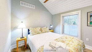 Une chambre à coucher comprenant un lit, une table de chevet, une lampe, un ventilateur de plafond, une fenêtre et une bouche d’aération fixée au mur.