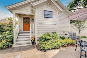 A house with a front porch, steps leading to a door, a small garden with bushes, and a patio area with a table and chairs.