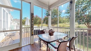 Un patio techado con una mesa de vidrio y sillas de mimbre, con vista a una zona residencial con una valla blanca y árboles.