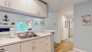 A kitchen with white cabinets, a sink, and a window. There are framed pictures on the wall and a blue crab poster.
