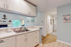 A kitchen with white cabinets, a sink, and a window. There are framed pictures on the wall and a blue crab poster. - Sea Colony condo with pool, hot tub, sauna, fitness room, gas fireplace & deck (Bethany Beach)