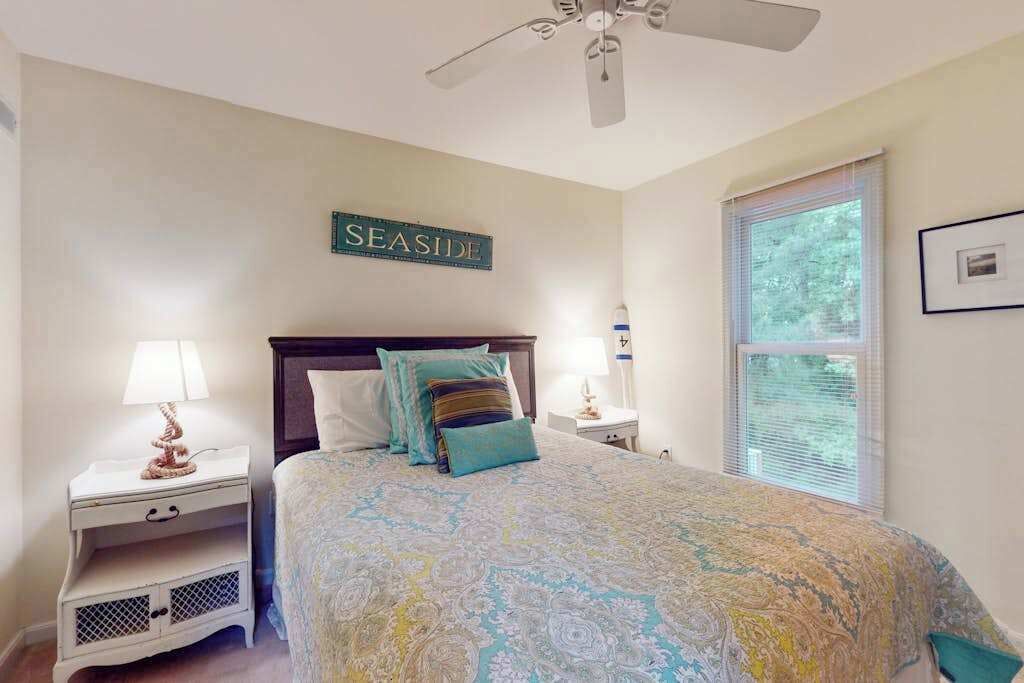 A bedroom with a bed, nightstands, a ceiling fan, a window with blinds, and a wall sign.