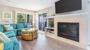 A living room with a sofa, a round table, a fireplace, and a TV mounted on the wall. - Sea Colony Tennis 1st-floor condo with gym, tennis court, & sauna (Bethany Beach)