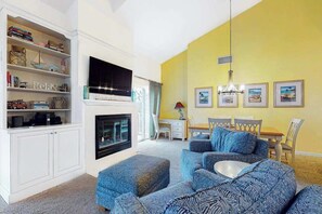 A living room with a fireplace, a television, a dining area with chairs, and a bookshelf with various items. - 2nd-floor Sea Colony Tennis condo with gym & sauna (Bethany Beach)