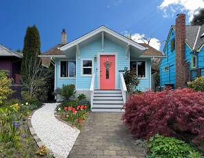 Ein Haus mit roter Tür, einer Veranda und einem Garten mit verschiedenen Pflanzen und Blumen.