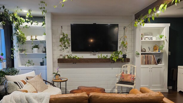 Ein Wohnzimmer mit einer Couch, einem an der Wand montierten Fernseher und Regalen mit Pflanzen und Büchern.