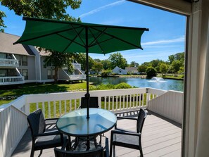 A deck with a glass table and chairs, a green umbrella, and a view of a lake and houses. - Sea Colony townhouse with Tennis, sauna, basketball court, & pool (Bethany Beach)