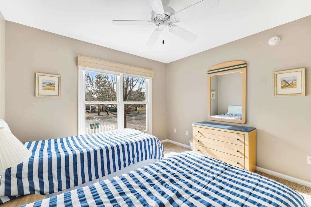 A bedroom with two beds, a dresser, a ceiling fan, and a window with blinds.