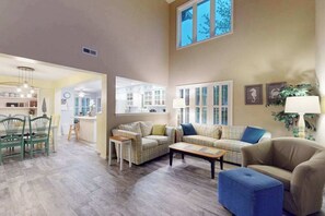 A living room with a sofa, chairs, a coffee table, and a lamp. There is a window with blinds and a plant on the wall. - Sea Colony townhome with tennis court, pool, gym, & basketball court (Bethany Beach)