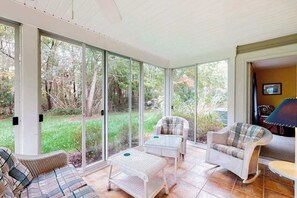 A sunlit room with a sofa, two armchairs, a small table, and a glass door leading to a garden. - Sea Colony Tennis townhouse with fireplace, sauna, pool, & basketball court (Bethany Beach)