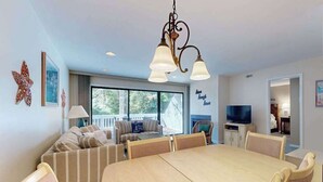 Uma sala de jantar com uma mesa grande e cadeiras, um conjunto de sofás, uma TV e um candelabro.