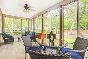Eine überdachte Terrasse mit Korbmöbeln, einem Deckenventilator und großen Fenstern mit Blick ins Freie.