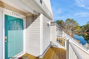 Une maison avec une porte de couleur turquoise, un revêtement extérieur blanc et une terrasse en bois dotée d’une balustrade blanche.