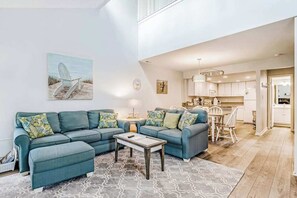 A modern living room with a sectional sofa, a coffee table, and a view into the kitchen. - Sea Colony Tennis 2 story 2 BR & loft townhome with sauna & pool (Bethany Beach)
