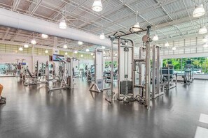 Un gimnasio espacioso con diversas máquinas de ejercicio, techo alto y ventanas grandes.
