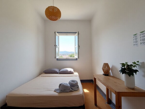 A bedroom with a single bed, a window with an open view, a wooden bedside table with a plant, and a hanging light fixture. - Santos (Lisbon)