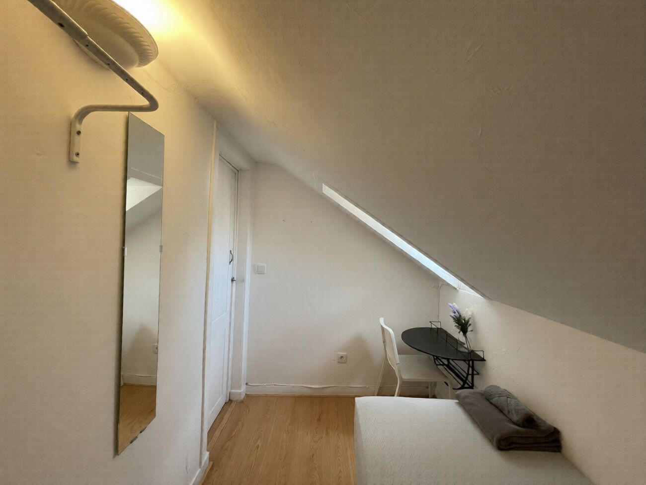 A small, narrow room with a sloped ceiling, a white chair, a small table, and a mirror on the wall.