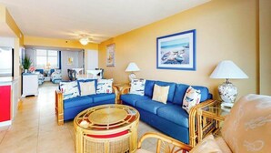 A living room with a blue sofa, wicker chairs, a glass coffee table, and a kitchen in the background. - 8th floor Sea Colony Oceancondo w/ balcony, elevator, and gym (Bethany Beach)