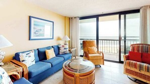 A living room with a blue sofa, wicker armchairs, a glass coffee table, and a large window with a view. - 8th floor Sea Colony Oceancondo w/ balcony, elevator, and gym (Bethany Beach)