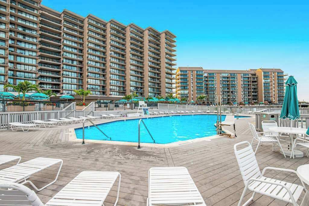 A hotel pool area with multiple lounge chairs, a swimming pool, and high-rise buildings in the background.