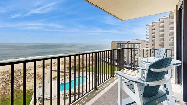 A balcony with a view of the beach and ocean, a pool, and a table with chairs. - Sea Colony Ocean 9th floor condo w/ elevator, tennis court, and pool (Bethany Beach)