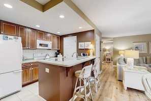 A kitchen with wooden cabinets, a white refrigerator, a microwave, and a central island with bar stools. - Sea Colony Tennis 1st floor condo w/ gym, basketball court, and shared sauna (Bethany Beach)