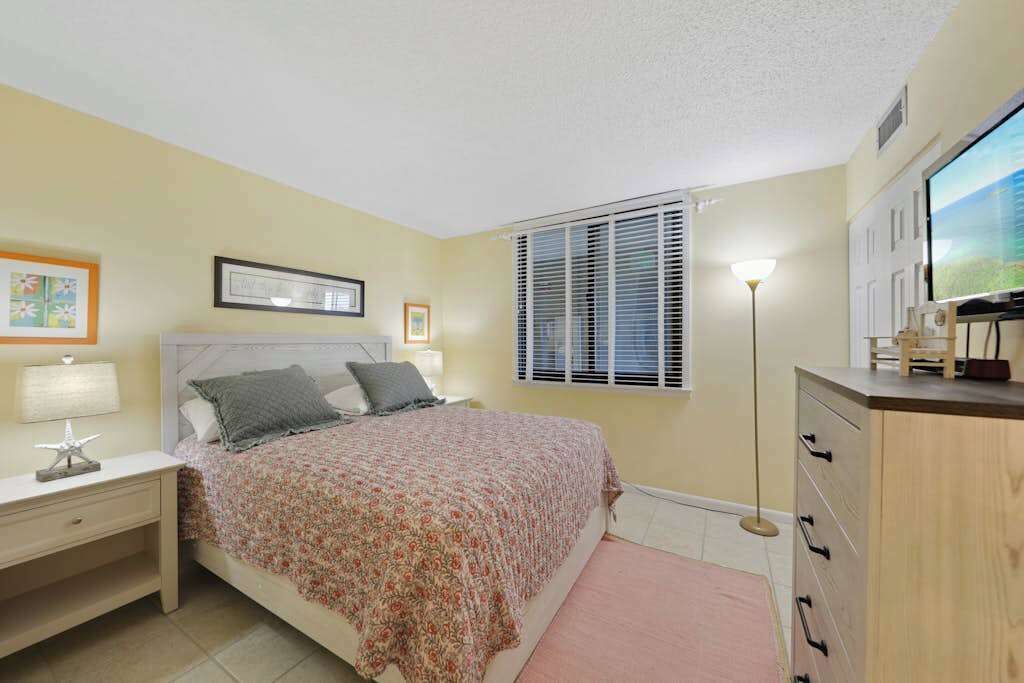 A bedroom with a bed, bedside table, lamp, framed picture, window with blinds, and a TV on a cabinet.