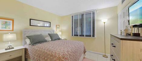 A bedroom with a bed, bedside table, lamp, framed picture, window with blinds, and a TV on a cabinet.