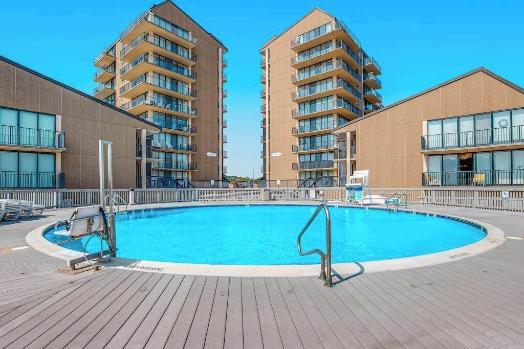 Une piscine entourée de deux immeubles de grande hauteur dotés de balcons, d’une terrasse en bois et surplombant un ciel bleu azur.