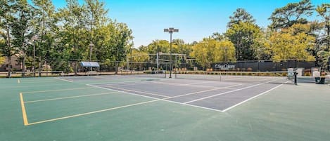 Cancha de tenis exterior con red, rodeada de árboles y una valla.