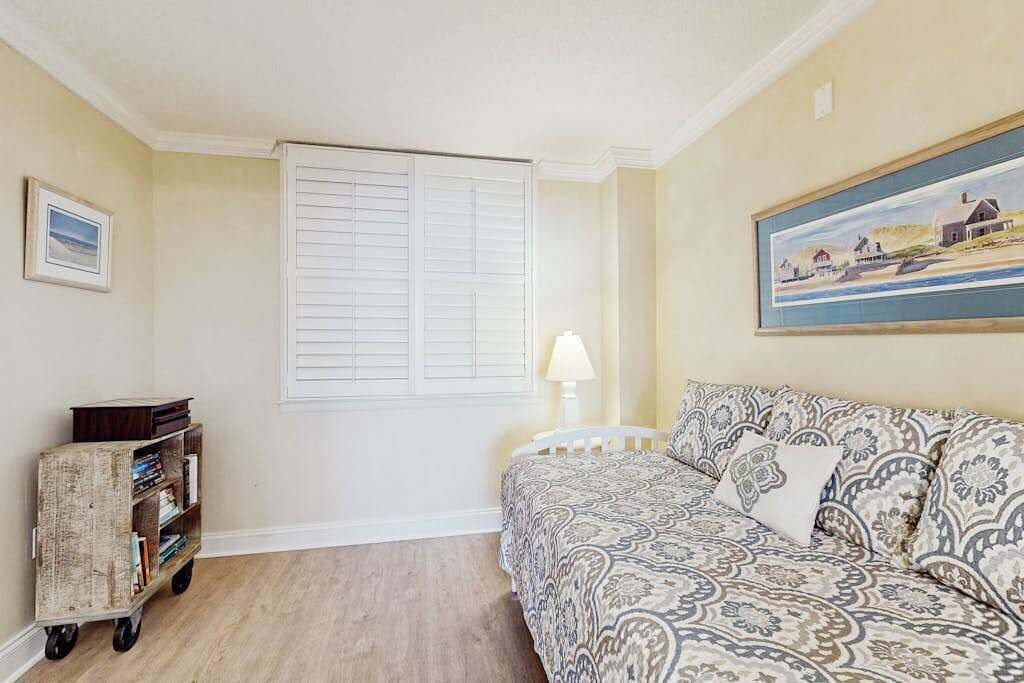 A room with a patterned sofa, a small cart with books, a window with shutters, and a painting on the wall.