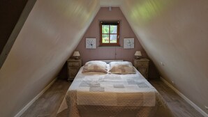 Ein Schlafzimmer mit schrägverdeckter Decke, einem Einzelbett mit Quiltdecke, zwei Nachttischen mit Lampen und einem Fenster mit Blick auf Grünflächen.