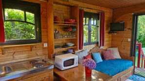 A cozy cabin interior with a kitchenette, a bed with pillows, a TV mounted on the wall, and a window with curtains. - Bohemian Caravan Cottage for 2–4 Guests near Honfleur with Pool (Fatouville-Grestain)