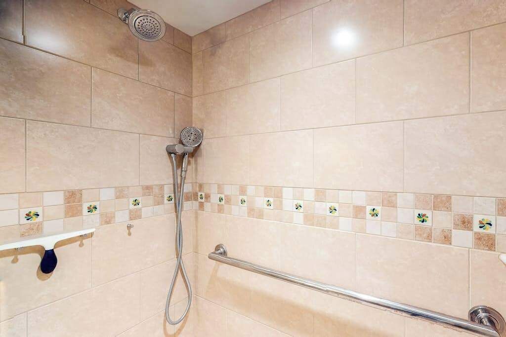 Un espace douche équipé d’un pommeau de douche à main, d’une barre d’appui et d’un mur carrelé agrémenté d’une bordure décorative.