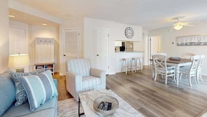 A living room with a sofa, a dining table with chairs, a kitchen area, and a clock on the wall. - Sea Colony Tennis 1st-floor condo with gym, tennis court & private beach (Bethany Beach)
