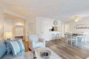 A living room with a sofa, a dining table with chairs, a kitchen area, and a clock on the wall. - Sea Colony Tennis 1st-floor condo with gym, tennis court & private beach (Bethany Beach)