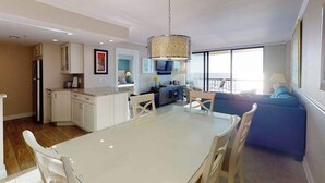 A modern kitchen with white cabinets, a dining table with chairs, a blue sofa, and a large window providing natural light. - Sea Colony Ocean 5th floor condo w/ balcony, shared sauna, and pool (Bethany Beach)
