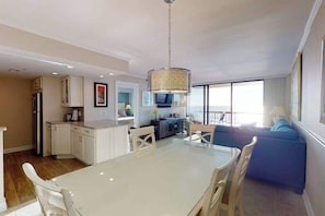 A modern kitchen with white cabinets, a dining table with chairs, a blue sofa, and a large window providing natural light. - Sea Colony Ocean 5th floor condo w/ balcony, shared sauna, and pool (Bethany Beach)