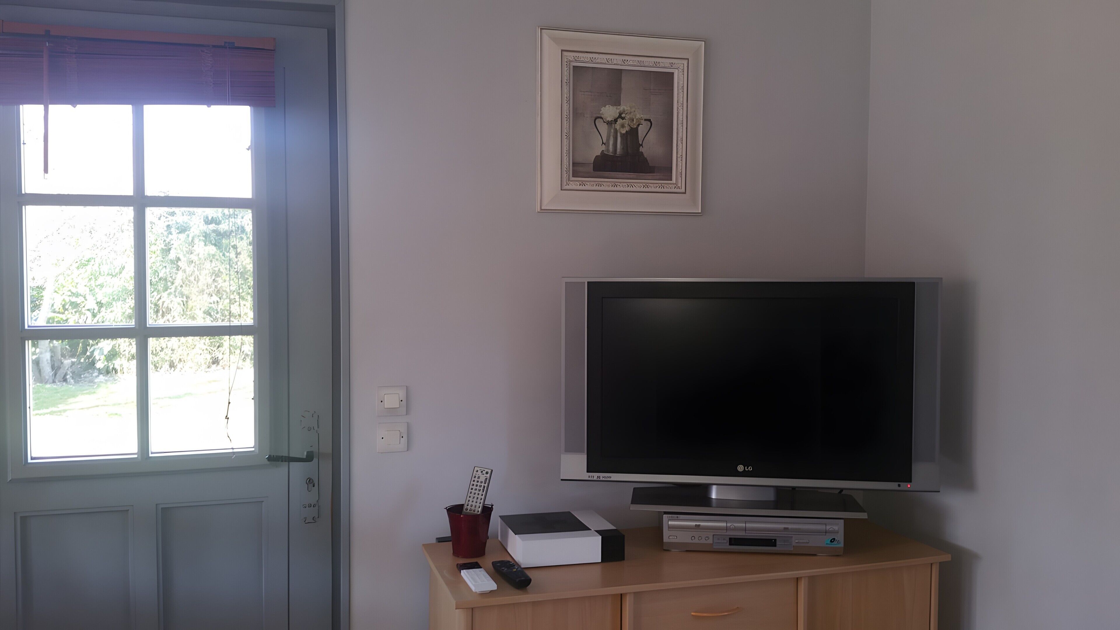 Huoneessa on televisio puisen kaapin päällä, seinällä on kehystetty taulu ja ikkunassa on sälekaihtimet.