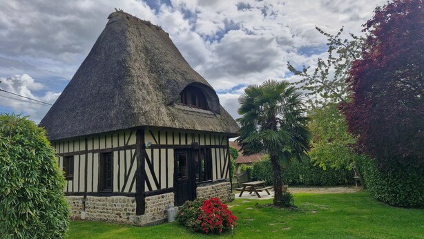 Una casa tradicional con techo de paja y base de piedra, rodeada de un jardín cuidado con una banca y una palmera.