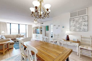 A spacious living area with a wooden dining table, white chairs, a chandelier, and a large window with curtains. - Sea Colony 3rd floor condo w/ free WiFi, tennis court, and shared sauna (Bethany Beach)
