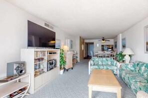 A living room with a TV, bookshelf, and a floral sofa. - Sea Colony 4th floor condo - shared pool, basketball court, partial ocean view (Bethany Beach)
