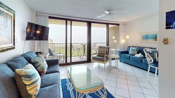 Ein modernes Wohnzimmer mit einer Couch, einem Couchtisch mit Glasplatte, einem Fernseher und einem Balkon mit Stühlen.