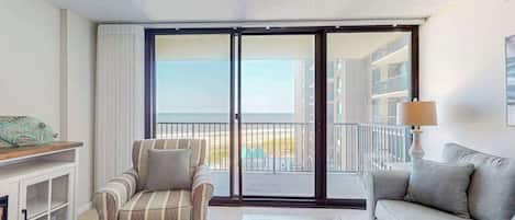 A hotel room with a sofa, armchair, and a view of the beach through a sliding door.