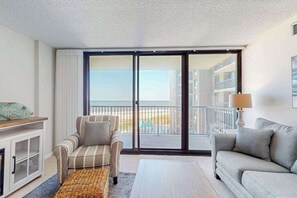 Habitación de hotel con sofá, sillón y vista a la playa a través de una puerta corrediza.