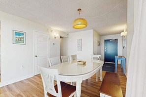 A dining area with a glass-top table, white chairs, a bench, a wicker pendant light, and a blue door in the background. - Sea Colony oceanfront condo with pool, hot tub, sauna, basketball, elevator (Bethany Beach)