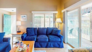 A living room with a blue sofa, a wooden coffee table, and a view through a sliding glass door to an outdoor deck. - Sea Colony Tennis 2nd-floor condo with basketball court, pool, & tennis court (Bethany Beach)